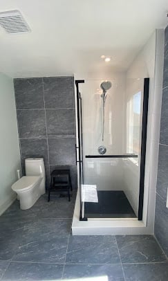 Modern bathroom with gray tile flooring, walk-in shower with glass door, white toilet, and showerhead mounted on tiled wall