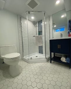 Modern bathroom with white hexagonal tile flooring, glass walk-in shower, white toilet, and dark vanity with mirror