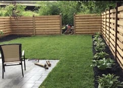 A modern backyard with green lawn, wooden fence panels, concrete patio, black chair, and planted garden beds
