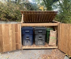 Wooden storage shed with open double doors revealing two black trash bins inside